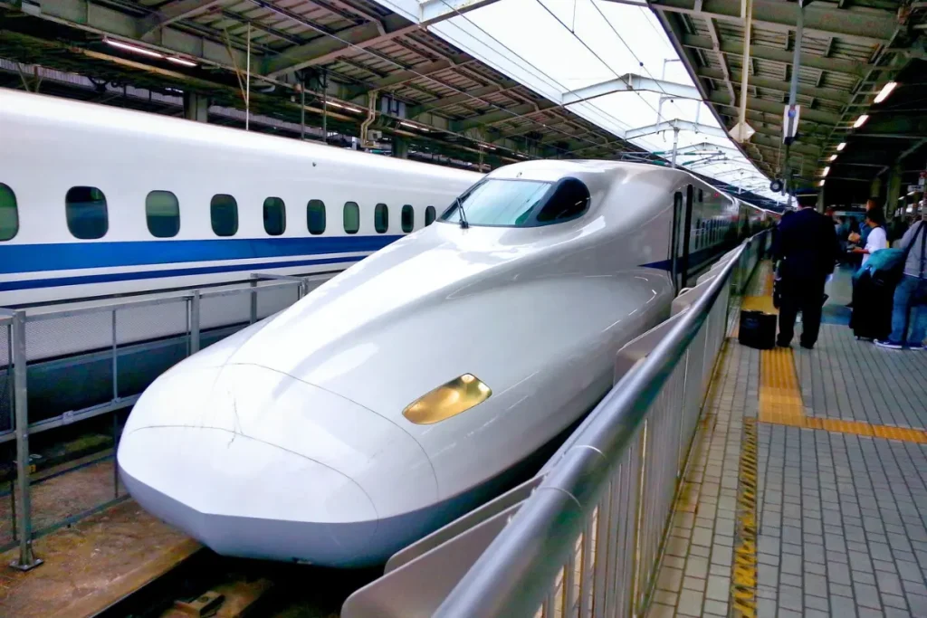 Train à grande vitesse Shinkansen blanc à quai dans une gare japonaise, avec voyageurs sur le quai et ambiance moderne typique des transports au Japon.