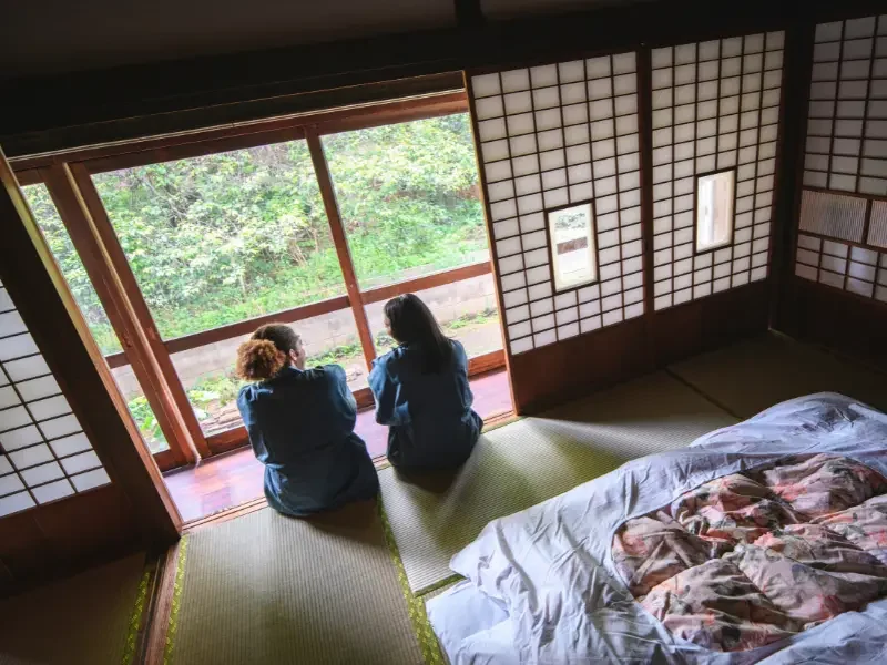 Chambre traditionnelle de ryokan avec vue sur jardin japonais Voyageuses assises sur des tatamis dans une chambre de ryokan traditionnel japonais, face à un jardin paisible.