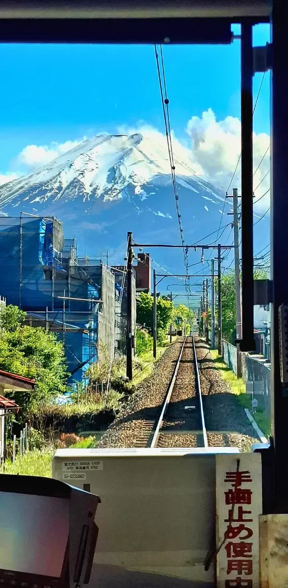 Vue depuis un train local dans la région des Fuji Five Lakes avec les rails menant vers le Mont Fuji enneigé sous un ciel bleu
