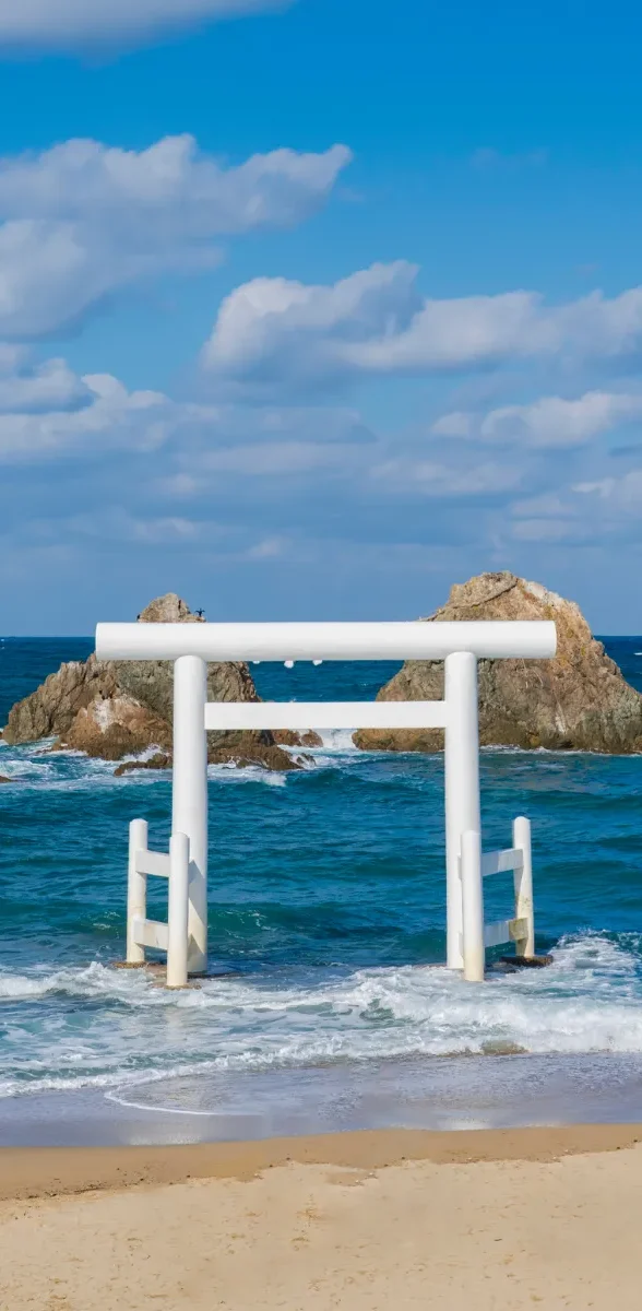 Torii blanc face à la mer sur la côte de Fukuoka au Japon, entre plage de sable clair, eau turquoise et ciel bleu lumineux.