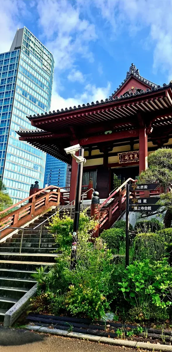 Temple traditionnel et architecture moderne à Tokyo Temple japonais traditionnel entouré de verdure à Tokyo, avec un immeuble moderne en verre en arrière-plan sous un ciel bleu lumineux.